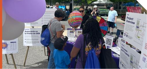Community members visit a booth at a Ciñco de Mayo celebration. The booth displays public engagement materials for the Peña NEPA and Design Project, including project information boards, maps, handouts, a QR code sign, and a colorful prize wheel. Several attendees reviewed materials and spoke with project representatives. Decorative balloons and event activities are visible around the booth in an outdoor festival setting.