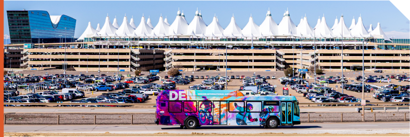 A colorful DEN-branded shuttle bus travels along a roadway at DEN with the the airport’s distinctive white peaked terminal roof behind the shuttle. The Westin Hotel and economy parking lots are also shown. 